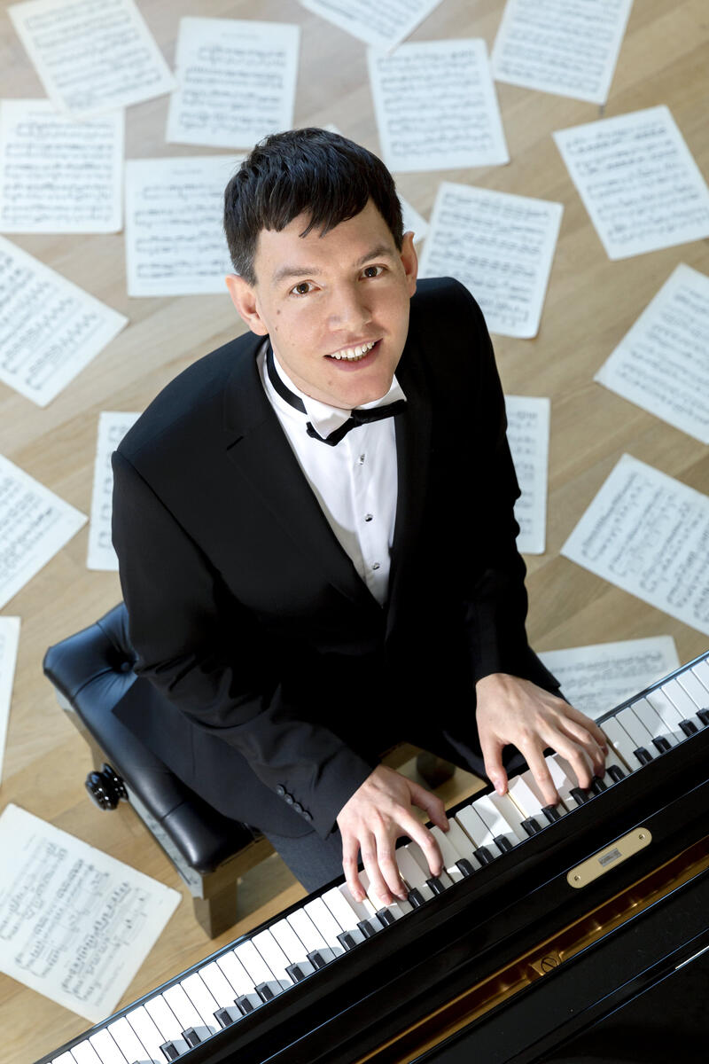 Matthew Shiel plays a Steinway grand piano whilst sheet music is strewn around him dramatically